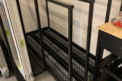 Trzy czarne metalowe konstrukcje ramowe ze wzorzystymi podporami u dołu ustawione pionowo przy ścianie w sali dydaktycznej montażu okien, obok przedmiotów w białych ramach i czarnego wózka z drewnianym blatem, wszystkie na szarej podłodze. projekt i nadzór Tatjana Kobuszewska trksystem.com