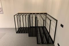 Czarny metalowy stelaż z wieloma pionowymi otworami stoi w rogu sali dydaktycznej montażu okien, jego solidna konstrukcja idealnie nadaje się do przechowywania dużych płaskich przedmiotów. Jasne ściany i szara wykładzina dywanowa zapewniają neutralne tło. projekt i nadzór Tatjana Kobuszewska trksystem.com