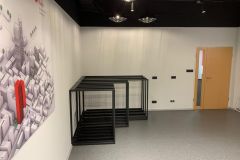 Sala szkoleniowa z białymi ścianami, zamkniętymi drewnianymi drzwiami, trzema pustymi czarnymi metalowymi stojakami i dużym plakatem ściennym przedstawiającym mapę miasta z wyróżnionym czerwonym budynkiem. Sufit ma kilka okrągłych opraw oświetleniowych i zabudowa okna. projekt i nadzór Tatjana Kobuszewska trksystem.com
