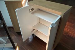 Nowoczesna drewniana szafka z białymi drzwiczkami jest otwarta, odsłaniając dwie półki w środku. Szafka stoi na podłodze z ciemnego drewna i jest oświetlona ciepłym światłem. Na ścianie w tle widoczne są zielone liście. projekt i nadzór Tatjana Kobuszewska trksystem.com