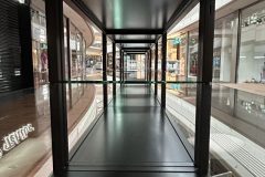 Symetryczny widok z wnętrza błyszczącej, pustej prostokątnej półki lub gabloty w centrum handlowym; fronty sklepów, jasne światła i odbicia są widoczne na wypolerowanych powierzchniach na zewnątrz gabloty. projekt i nadzór Tatjana Kobuszewska trksystem.com