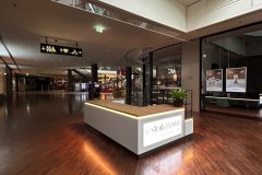 Nowoczesne wnętrze centrum handlowego w galerii Manufaktura z wypolerowanymi drewnianymi podłogami, zakrzywionymi balustradami nad nimi i białą wyspą handlową z napisem Słoik Home w centrum. Sklepy ze szklanymi frontami i podświetlanymi szyldami wypełniają cichą, pustą przestrzeń. projekt i nadzór Tatjana Kobuszewska trksystem.com