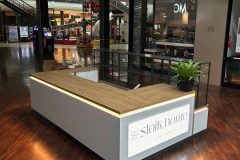 Mały, nowoczesny kiosk z drewnianymi i białymi panelami znajduje się w centrum handlowym. Na jego świecącym szyldzie widnieje napis Stolk Home. Jeden z rogów zdobi zielona roślina doniczkowa. Centrum handlowe jest ciche, a w tle widać sklepy, miejsca do siedzenia i automaty z napojami. projekt i nadzór Tatjana Kobuszewska trksystem.com