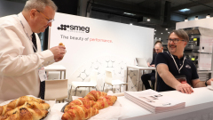 Dwóch mężczyzn wchodzi w interakcję na stoisku targowym z profesjonalnym znakiem smeg za nimi. Jeden z nich próbuje rogalika z tacy, podczas gdy drugi, siedzący przy ladzie z broszurami, uśmiecha się. W tle inna osoba pracuje na laptopie. projekt i nadzór Tatjana Kobuszewska trksystem.com