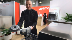 Mężczyzna w czarnej koszuli trzyma części zmywarki, stojąc w nowoczesnym salonie wystawowym. Dwie osoby rozmawiają przy biurku w tle obok znaku Smeg Professional. Widoczne są zielone rośliny i urządzenia kuchenne ze stali nierdzewnej. projekt i nadzór Tatjana Kobuszewska trksystem.com