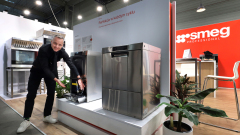 Uśmiechnięty mężczyzna w czerni otwiera zmywarkę ze stali nierdzewnej w nowoczesnym salonie AGD. Obok niego znajduje się kolejna zmywarka; w pobliżu zielone rośliny i biurko. W tle widoczna jest czerwona ściana z białym logo "smeg PROFESSIONAL". projekt i nadzór Tatjana Kobuszewska trksystem.com