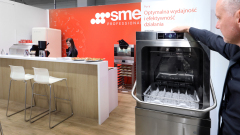 Mężczyzna demonstruje komercyjną zmywarkę obok lady ze stołkami barowymi, gdzie kobieta przygotowuje jedzenie. W tle znajduje się jasnoczerwone stoisko SME Professional z polskim tekstem promującym optymalną wydajność i wydajność. projekt i nadzór Tatjana Kobuszewska trksystem.com