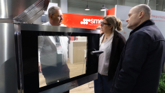 Trzy dorosłe osoby stoją przed dużym piekarnikiem ze stali nierdzewnej na stoisku wystawowym. Jeden z mężczyzn, ubrany w białą koszulę i krawat, objaśnia urządzenie mężczyźnie i kobiecie, oboje w ciemnych ubraniach wierzchnich. Na stoisku znajduje się logo SMEG i nowoczesne, profesjonalne otoczenie. projekt i nadzór Tatjana Kobuszewska trksystem.com