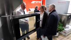 Trzy dorosłe osoby stoją wokół dużego piekarnika ze stali nierdzewnej na targach. Mężczyzna w białej koszuli gestykuluje do piekarnika, rozmawiając z kobietą i mężczyzną. W tle widoczne są czerwone i białe stoiska z tekstem i roślinami. projekt i nadzór Tatjana Kobuszewska trksystem.com