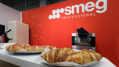 Dwie tace z rogalikami stoją na ladzie w st6oisku targowym smeg, przed czerwoną ścianą z białym logo "smeg PROFESSIONAL". W tle znajduje się kremowa lodówka i srebrny piekarnik. Rogaliki wyglądają na świeże i złocistobrązowe. projekt i nadzór Tatjana Kobuszewska trksystem.com