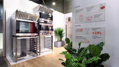 Stoisko Smeg Nowoczesna komercyjna kuchnia z dużymi piekarnikami ze stali nierdzewnej, stojakami i okapem wentylacyjnym na drewnianej podłodze. Dwa plakaty informacyjne z wykresami i statystykami w języku polskim znajdują się na białej ścianie obok zielonych roślin doniczkowych. projekt i nadzór Tatjana Kobuszewska trksystem.com
