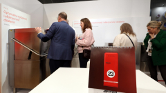 Czerwony kalendarz biurkowy stoi na białym stole na pierwszym planie. W tle cztery dorosłe osoby stoją przy kołowrotku ze stali nierdzewnej w dobrze oświetlonym stoisku wystawowym z białymi ścianami i polskim tekstem o wydajności i bezpieczeństwie. projekt i nadzór Tatjana Kobuszewska trksystem.com