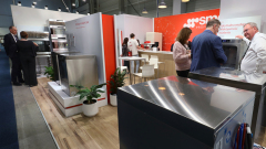 Ludzie wchodzą w interakcje wokół nowoczesnych urządzeń kuchennych na stoisku targowym. Widoczny jest biało-czerwony wystrój, rośliny doniczkowe i lady wystawowe. Dwie pary rozmawiają o urządzeniach, podczas gdy inna osoba stoi w pobliżu maszyny ze stali nierdzewnej. projekt i nadzór Tatjana Kobuszewska trksystem.com