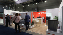 Ludzie w strojach biznesowych wchodzą w interakcje na stoisku targowym prezentującym profesjonalne urządzenia kuchenne SMEG. Stoisko ma czerwono-biały branding, nowoczesne piekarniki, zmywarki i lady informacyjne na jasnej drewnianej podłodze w przemysłowym oświetleniu. projekt i nadzór Tatjana Kobuszewska trksystem.com