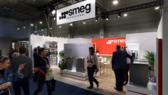 Stoisko wystawowe Smeg Professional prezentuje urządzenia takie jak zmywarki i piekarniki. Kilka osób przechodzi obok lub wchodzi w interakcję z personelem, podczas gdy duże logo Smeg wisi nad i na ścianach. Stoisko ma czerwono-biały branding w dobrze oświetlonej hali eventowej. projekt i nadzór Tatjana Kobuszewska trksystem.com