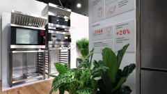 Nowoczesna witryna kuchenna zawiera piekarniki i urządzenia ze stali nierdzewnej, zielone rośliny doniczkowe, drewnianą podłogę i ścianę z grafiką informacyjną i tekstem w języku polskim na temat wydajności i wydajności piekarnika. Otoczenie wydaje się jasne i czyste. projekt i nadzór Tatjana Kobuszewska trksystem.com