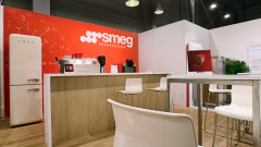 W nowoczesnym salonie wystawowym znajduje się czerwono-biała ekspozycja "Smeg Professional" z lodówką w stylu retro, ekspresami do kawy i eleganckimi białymi krzesłami wokół jasnych drewnianych stołów na drewnianej podłodze, pod industrialnym oświetleniem. projekt i nadzór Tatjana Kobuszewska trksystem.com