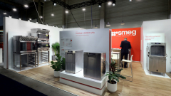 Nowoczesne stoisko targowe Smeg Professional prezentuje duże urządzenia kuchenne ze stali nierdzewnej. Mężczyzna w garniturze stoi przy białej ladzie z krzesłami. Stoisko ma czerwono-biały branding, rośliny doniczkowe i drewnianą podłogę pod jasnymi lampami sufitowymi. projekt i nadzór Tatjana Kobuszewska trksystem.com