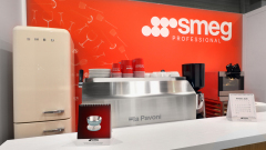 Ekspozycja SMEG Professional obejmuje kremową lodówkę retro, ekspres do kawy La Pavoni, czerwone kubki SMEG i kalendarz na białej ladzie. Czerwona ściana tła ma branding SMEG i kontury szklanych naczyń. projekt stoiska i nadzór Tatjana Kobuszewska trksystem.com