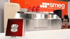 Ekspres do kawy la Pavoni ze stali nierdzewnej stoi na blacie z ułożonymi talerzami i czerwonymi filiżankami SMEG. W tle znajduje się lodówka SMEG. Czerwono-biały baner SMEG Professional jest wyświetlany z tyłu, a broszura produktu stoi na pierwszym planie. projekt i nadzór Tatjana Kobuszewska trksystem.com