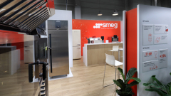 Nowoczesny showroom z profesjonalnymi urządzeniami Smeg, w tym lodówkami i piekarnikami, wyeksponowanymi na podłodze z jasnego drewna. W tle znajduje się pomarańczowa ściana z marką Smeg, lada z białymi stołkami, rośliny doniczkowe i oznakowanie informacyjne. projekt i nadzór Tatjana Kobuszewska trksystem.com