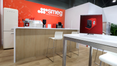 Nowoczesny showroom z drewnianą ladą z białymi stołkami, białą lodówką i ekspresami do kawy. Ściana jest jaskrawoczerwona z logo SMEG PROFESSIONAL. Na stoliku z przodu znajduje się kalendarz biurkowy z czerwono-czarnym urządzeniem SMEG. projekt i nadzór Tatjana Kobuszewska trksystem.com