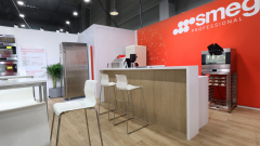 Nowoczesny salon wystawowy wyposażony jest w profesjonalne urządzenia kuchenne Smeg, w tym lodówkę i piekarnik, z czerwoną ścianą akcentującą. Białe krzesła stoją wzdłuż drewnianej lady na jasnej drewnianej podłodze, a jasne oświetlenie podkreśla czystą, nowoczesną przestrzeń. projekt i nadzór Tatjana Kobuszewska trksystem.com