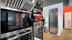 Salon wystawowy prezentuje nowoczesne urządzenia kuchenne ze stali nierdzewnej, w tym piekarniki, płyty grzewcze i lodówki. Urządzenia są starannie rozmieszczone z jasnym oświetleniem, a w tle widoczna jest zielona roślina i czerwona ściana akcentująca. projekt i nadzór Tatjana Kobuszewska trksystem.com