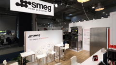 Nowoczesne stoisko targowe dla Smeg Professional prezentuje piekarniki, lodówkę ze stali nierdzewnej, białe stoły z krzesłami i ścianki informacyjne. czysty, minimalistyczny design. projekt i nadzór Tatjana Kobuszewska trksystem.com