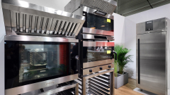 Ekspozycja nowoczesnych komercyjnych urządzeń kuchennych ze stali nierdzewnej, w tym dwóch dużych piekarników, okapu kuchennego i wysokiej lodówki, ustawionych na jasnej ścianie z drewnianą podłogą i zieloną rośliną doniczkową w tle. projekt i nadzór Tatjana Kobuszewska trksystem.com