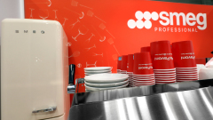 Kremowa lodówka SMEG stoi obok ułożonych w stos białych talerzy i czerwonych filiżanek do kawy na blacie ze stali nierdzewnej. Tło stanowi czerwona ściana z logo SMEG Professional i białymi rysunkami naczyń kuchennych. projekt i nadzór Tatjana Kobuszewska trksystem.com