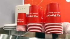 Stos czerwonych papierowych kubków do kawy z białymi brzegami, z napisem "La Pavoni" do góry nogami, leży na stosie białych talerzy na blacie. W tle widoczne jest urządzenie Smeg w stylu retro i czerwona ściana. projekt i nadzór Tatjana Kobuszewska trksystem.com