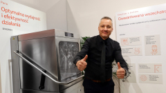 Uśmiechnięty mężczyzna w czarnej koszuli i krawacie stoi obok komercyjnej zmywarki, trzymając dwa kciuki w górę. Za nim znajdują się znaki w języku polskim z tekstem o wydajności i niezawodności. Wygląda to na targi lub prezentację produktu. projekt i nadzór Tatjana Kobuszewska trksystem.com