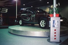 Bordowy SUV z brandingiem JVC jest wyświetlany na podniesionej, okrągłej, metalowej platformie na wystawie wewnętrznej, tworząc centralny element muzycznego stoiska JVC. Na pierwszym planie znajduje się stojak na broszury z czerwonymi przyciskami i dwie rośliny doniczkowe. projekt i nadzór Tatjana Kobuszewska trksystem.com