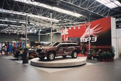 Bordowy SUV z brandingiem JVC jest prezentowany na okrągłej platformie podczas targów motoryzacyjnych, tworząc centralny element muzycznego stoiska JVC. Nieopodal, inny markowy samochód stoi przed odważnym czerwono-czarnym tłem JVC MP3. projekt i nadzór Tatjana Kobuszewska trksystem.com