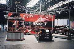 Muzyczne stoisko JVC zawiera samochody, stoiska promocyjne i duży wyświetlacz MP3 z wyciągniętą ręką. Stoisko jest jasno oświetlone górnymi światłami, czerwonym wystrojem i banerami tworzącymi dynamiczną atmosferę. projekt i nadzór Tatjana Kobuszewska trksystem.com