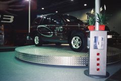 Ciemny SUV z brandingiem JVC jest wystawiony na okrągłej, podniesionej metalowej platformie podczas imprezy wewnętrznej, stanowiącej część muzycznego stoiska JVC. Na pobliskim stoisku znajduje się broszura i dwie małe rośliny doniczkowe. Tło jest słabo oświetlone. projekt i nadzór Tatjana Kobuszewska trksystem.com