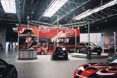 Na wystawie samochodowej w dużej hali znajduje się kilka samochodów, czerwono-czarne muzyczne stoisko JVC i duży czerwony baner z MP3 i obrazami rąk używających sprzętu audio. Jasne światła nad głową oświetlają scenę. projekt i nadzór Tatjana Kobuszewska trksystem.com