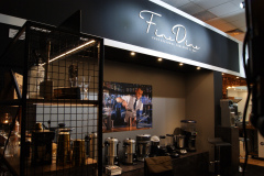 Nowoczesne stoisko wystawowe dla "FineDine" z dużym znakiem, różnymi profesjonalnymi naczyniami do napojów i sprzętem do kawy. Zdjęcie barmana mieszającego drinka jest zamontowane na ścianie, a półki prezentują szkło i narzędzia barowe w ciepłym oświetleniu. projekt i nadzór Tatjana Kobuszewska trksystem.com