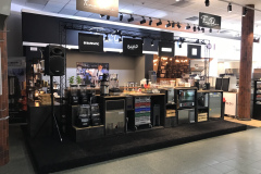 Dobrze oświetlona lada kawiarni prezentuje ekspresy do kawy, blendery, kubki i składniki. Nad nimi widnieją napisy Fine Dine, BUNN i BACKUP. Na półkach znajdują się materiały eksploatacyjne, a na ekranie wyświetlany jest obraz o tematyce kawowej. Konfiguracja znajduje się na podniesionej czarnej platformie w przestronnym pomieszczeniu. projekt i nadzór Tatjana Kobuszewska trksystem.com