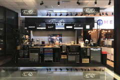 Nowoczesny stojak na kawę z różnym sprzętem, filiżankami i materiałami eksploatacyjnymi. Powyżej wiszą czarne szyldy FineDine. Na ekranie telewizora wyświetlany jest szef kuchni, a także tablica z menu. Ciepłe oświetlenie i stylowa, zorganizowana aranżacja są widoczne w otoczeniu centrum handlowego. projekt i nadzór Tatjana Kobuszewska trksystem.com