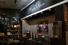 Nowoczesny bar kawowy o nazwie "Fine Dine" z czarno-białym szyldem, ekspresami do kawy, filiżankami i sprzętem do parzenia na ciemnej ladzie. Podobnie jak stylowe stoisko targowe, półki z zapasami i duże zdjęcie baristy dodają przytulnego, słabo oświetlonego klimatu. projekt i nadzór Tatjana Kobuszewska trksystem.com