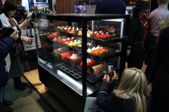 Grupa ludzi gromadzi się wokół szklanej gabloty wypełnionej kolorowymi wypiekami i deserami. Kilka osób robi zdjęcia swoimi telefonami, podczas gdy inni przyglądają się smakołykom. Wydaje się, że scena rozgrywa się podczas intensywnego wydarzenia lub wystawy. projekt i nadzór Tatjana Kobuszewska trksystem.com