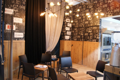 Nowoczesne wnętrze kawiarni z czarnymi krzesłami wokół małych stolików, drewnianymi panelami na ścianach i tapetą w stylu tablicy kredowej. Z sufitu zwisa ciepłe oświetlenie wiszące. Przezroczysta zasłona częściowo zakrywa ciemną draperię, a w tle widoczny jest sprzęt kuchenny ze stali nierdzewnej. projekt i nadzór Tatjana Kobuszewska trksystem.com