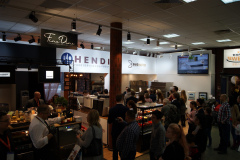 Zajęte stoisko targowe wewnątrz budynku zawiera ekspozycje sprzętu gastronomicznego, ludzi przeglądających i rozmawiających, podświetlane znaki firmowe i próbki produktów na ladach. Uczestnicy ustawiają się w kolejce i wchodzą w interakcje z personelem w jasnej, nowoczesnej atmosferze wystawienniczej. projekt i nadzór Tatjana Kobuszewska trksystem.com