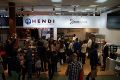 Na targach lub wystawie pod dachem znajdują się stoiska firm Hendi Food Service Equipment i BakeMaster. Wiele osób stoi, rozmawia i przegląda produkty i wyświetlacze, tworząc żywą atmosferę pod jasnymi światłami sufitowymi. projekt i nadzór Tatjana Kobuszewska trksystem.com