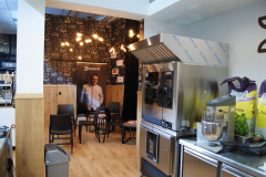 Nowoczesne wnętrze kawiarni z drewnianymi podłogami i ścianami, czarnymi krzesłami ustawionymi w małej części wypoczynkowej, ścianą pokrytą grafiką w stylu tablicy kredowej i różnymi urządzeniami kuchennymi, w tym dużym metalowym ekspresem do kawy i mikserem stojącym. W tle znajduje się plakat szefa kuchni. projekt i nadzór Tatjana Kobuszewska trksystem.com