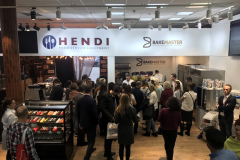 Tłum gromadzi się przy stoisku ze sprzętem gastronomicznym ze znakami Hendi i BakeMaster nad głową. Ludzie wchodzą w interakcje, próbują jedzenia i obserwują pokazy wypieków. Na stoisku znajdują się nowoczesne urządzenia kuchenne i szklana gablota z deserami pod jasnym oświetleniem. projekt i nadzór Tatjana Kobuszewska trksystem.com