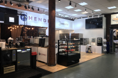 Nowoczesne stoisko targowe prezentuje sprzęt gastronomiczny Hendi. Na stoisku znajdują się urządzenia kuchenne, piekarniki, szklana gablota z ciastkami, ekspres do kawy i jasne oświetlenie. Znaki firmowe i duży telewizor z płaskim ekranem są widoczne nad czystą, zorganizowaną konfiguracją. projekt i nadzór Tatjana Kobuszewska trksystem.com