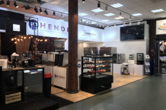 Nowoczesne stoisko targowe prezentuje sprzęt gastronomiczny Hendi i Bakemaster. Na stoisku znajdują się urządzenia kuchenne, szklana gablota z ciastkami, ekrany cyfrowe i jasne oświetlenie górne. Otoczenie jest czyste i uporządkowane, z widocznym oznakowaniem marki. projekt i nadzór Tatjana Kobuszewska trksystem.com